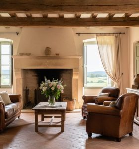 Sunlit French country living room with a weathered stone fireplace, hand-hewn oak beams, cream linen drapes, and heirloom pottery on a rustic sideboard