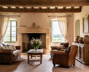 Sunlit French country living room with a weathered stone fireplace, hand-hewn oak beams, cream linen drapes, and heirloom pottery on a rustic sideboard